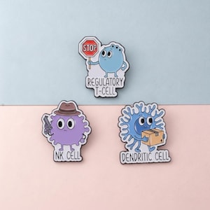 Könnte beinhalten: Drei Emaille-Anstecknadeln mit Cartoon-Designs von Immunzellen. Eine blaue Zelle hält ein Stoppschild mit der Aufschrift "Regulatory T-Cell". Eine lila Zelle mit Hut hält eine Pistole mit der Aufschrift "NK Cell". Eine blaue Zelle trägt eine Box mit der Aufschrift "Dendritic Cell".