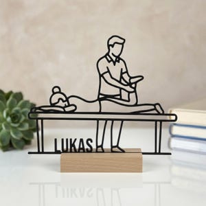 May include: A black metal wire-art sculpture depicting a person performing a massage on another individual lying on a table. The design includes the name LUKAS integrated into the frame, mounted on a rectangular light-coloured wooden base.