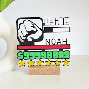 Könnte beinhalten: Dekoratives Schild mit einer schwarz-weißen Grafik einer Faust, dem Namen "NOAH" und dem Text "03:02". Das Schild hat einen roten Streifen, grünen Text mit der Zahl "999999999" und fünf gelbe Sterne. Es ist auf einem kleinen Holzsockel montiert.