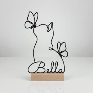 May include: A black wire sculpture featuring the silhouette of a cat with two butterflies and the name Bella written in cursive script, mounted on a rectangular light-colored wooden base. The design is created from a single continuous wire line.