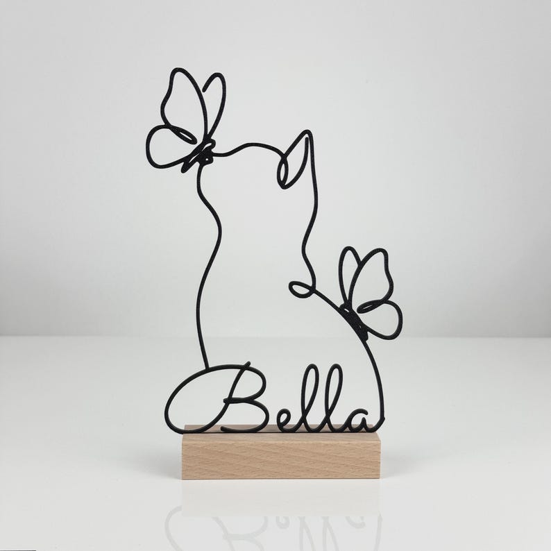 May include: A black wire sculpture featuring the silhouette of a cat with two butterflies and the name Bella written in cursive script, mounted on a rectangular light-colored wooden base. The design is created from a single continuous wire line.