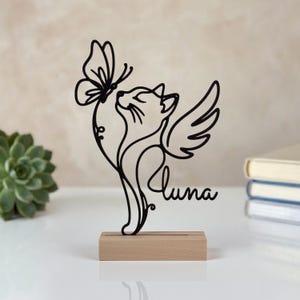 Könnte beinhalten: Eine schwarze Drahtkunst-Skulptur, die eine Katze mit Flügeln zeigt, die einen Schmetterling betrachtet, mit dem Namen Luna in Schreibschrift. Die Skulptur ist auf einem rechteckigen, hellen Holzsockel montiert und bildet ein durchgehendes Liniendesign.