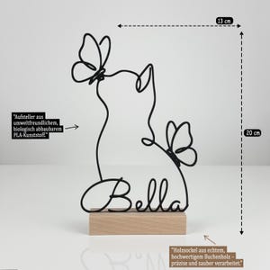 May include: A black wire sculpture of a cat with two butterflies and the name Bella, mounted on a rectangular wooden base. The sculpture measures 13 cm in width and 20 cm in height. The text indicates it is made of biodegradable PLA plastic.