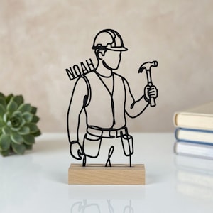 Op de afbeelding: Een zwart draadsculptuur van een bouwvakker met een veiligheidshelm en een vest met een gereedschapsgordel, die een hamer vasthoudt. De naam NOAH is in het ontwerp verwerkt. Het beeld staat op een rechthoekige, lichtgekleurde houten voet.