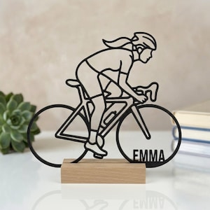 Puede incluir: Una silueta negra de un ciclista sobre una bicicleta de carretera, montada sobre una base rectangular de madera clara. El ciclista está inclinado hacia adelante en posición de carrera. El nombre EMMA está recortado en letras mayúsculas.