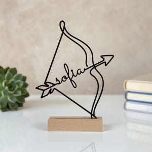 Può includere: Una scultura in filo metallico nero a forma di arco e freccia con il nome Sofia integrato nell'asta della freccia. La scultura è montata su una base rettangolare in legno chiaro, creando un oggetto decorativo autoportante.
