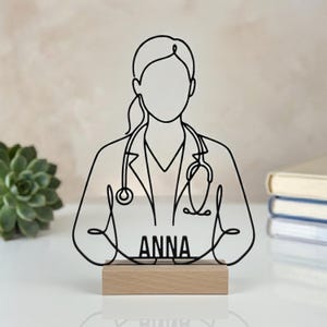 Op de afbeelding: Een zwarte draadkunstsculptuur van een arts met een doktersjas en een stethoscoop om de nek. De naam ANNA staat in zwarte hoofdletters op de borst. De sculptuur is bevestigd op een rechthoekige basis van lichtgekleurd hout.
