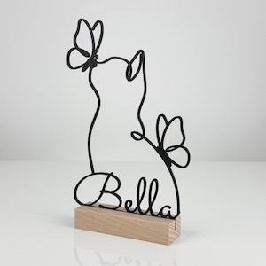 May include: A black wire sculpture featuring the outline of a cat silhouette with two butterflies and the name Bella in cursive script, mounted on a rectangular light-colored wooden base. The design is composed of continuous metal lines.