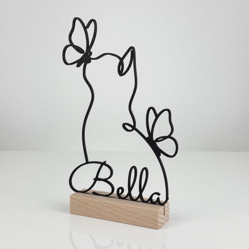 May include: A black wire sculpture featuring the outline of a cat silhouette with two butterflies and the name Bella in cursive script, mounted on a rectangular light-colored wooden base. The design is composed of continuous metal lines.