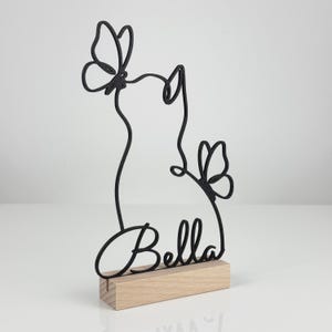 May include: A black wire-frame sculpture featuring the silhouette of a cat with two butterflies and the name Bella in cursive script, mounted on a rectangular light-colored wooden base. The design is linear and minimalist in its construction.
