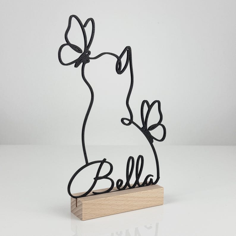 May include: A black wire-frame sculpture featuring the silhouette of a cat with two butterflies and the name Bella in cursive script, mounted on a rectangular light-colored wooden base. The design is linear and minimalist in its construction.