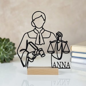 Könnte beinhalten: Eine schwarze Metallsilhouette einer Richterfigur in Robe, die in einer Hand einen Hammer und in der anderen eine Waage hält. Die Figur ist auf einem rechteckigen hellen Holzsockel montiert und zeigt den Namen ANNA als schwarze Metallschrift.