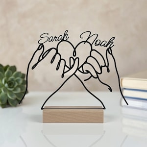 Op de afbeelding: Een zwart draadsculptuur met twee handen waarvan de pinken in elkaar zijn gehaakt om een hartvorm te vormen. De namen Sarah en Noah staan in cursief schrift boven de handen. Het sculptuur is bevestigd op een rechthoekige houten voet.
