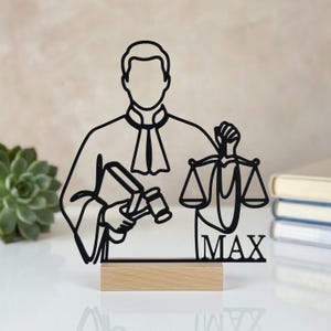 Può includere: Una sagoma in filo metallico nero di un giudice in toga, che tiene un martelletto in una mano e una bilancia della giustizia nell'altra. Il nome MAX è integrato nel design in basso a destra. La figura è montata su una base rettangolare in legno.