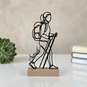 Peut inclure: Une sculpture en fil de métal noir représentant une personne en randonnée avec un sac à dos et des bâtons de marche, intégrant le prénom Sarah en écriture cursive, fixée sur un socle rectangulaire en bois clair.