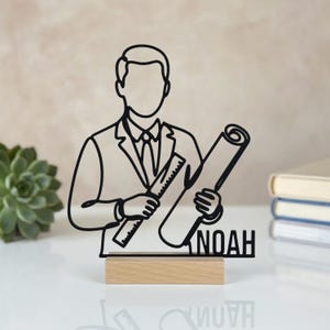 May include: A black wire-frame silhouette of an architect wearing a suit, holding a ruler and a rolled blueprint. The figure is mounted on a rectangular light-coloured wooden base. The name NOAH is displayed in black capital letters at the bottom.