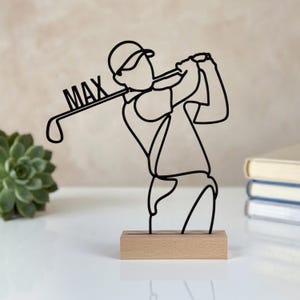 May include: A black wire sculpture depicting a golfer in a swing motion, mounted on a rectangular light wood base. The golf club shaft features the name MAX integrated into the design. The sculpture is crafted from a single continuous wire line.