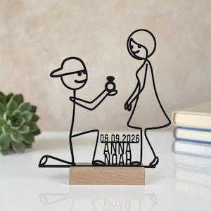 Può includere: Una sagoma in filo metallico nero che raffigura una persona in ginocchio che fa una proposta, montata su una base rettangolare in legno. Il design include il testo 06.09.2026, ANNA e NOAH integrato nella struttura tra le due figure.