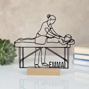 Könnte beinhalten: Eine schwarze Drahtkunstskulptur, die einen Massagetherapeuten bei der Massage eines auf einem Tisch liegenden Kunden darstellt. Die Skulptur ist auf einem rechteckigen hellen Holzsockel montiert und enthält den Namen EMMA im Drahtdesign.