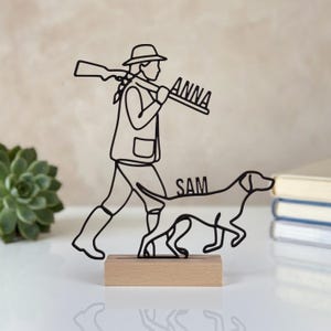 Può includere: Una scultura in filo metallico nero raffigurante una persona con cappello e stivali, che porta un fucile sulla spalla con il nome ANNA integrato nel design, che cammina accanto a un cane con il nome SAM integrato nel corpo, su una base in legno.