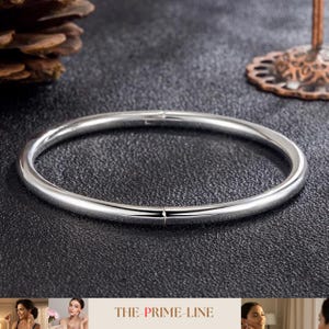 Könnte beinhalten: Ein silberfarbenes Armreif-Armband mit glattem, abgerundetem Design. Das Armband wird auf einer dunklen, strukturierten Oberfläche gezeigt. Der Text "THE PRIME-LINE" befindet sich am unteren Bildrand.
