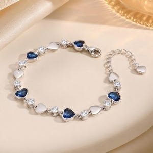 May include: A silver-toned bracelet featuring alternating heart-shaped blue crystals and clear round stones. The chain includes a lobster claw clasp and an adjustable extender chain finished with a small heart charm at the end of the link section.