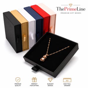 Puede incluir: Una colección de cajas de regalo rectangulares con cajón deslizante en negro, dorado, blanco, azul marino, rojo y rosa claro, cada una con una lengüeta de cinta. Un collar de oro rosa con un colgante texturizado descansa en una caja negra abierta.