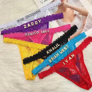 Könnte beinhalten: Eine Kollektion aus sechs Spitzen-Tangas in Weiß, Lila, Pink, Schwarz, Blau und Rot. Jedes Stück hat einen dekorativen Bund mit Strassbuchstaben, die MRS EDGEMON, ZADDY, MEXICO 2024, KHALIL, BRIAN WEST und IVAN buchstabieren.