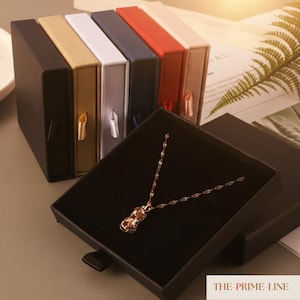 May include: A gold necklace with a pendant rests in an open black jewelry box. Several closed boxes in various colors, including black, gold, white, navy, red, and beige, are arranged behind it. Each box has a ribbon pull.