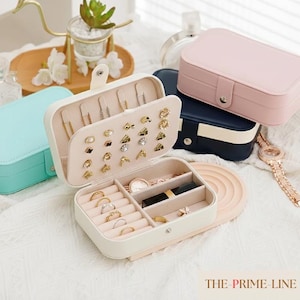 Puede incluir: Caja de joyería crema abierta, con múltiples compartimentos, que muestra anillos, collares y aretes. Se ven otras cajas de joyería cerradas en azul claro, azul marino y rosa. También hay un reloj dorado y lápiz labial.