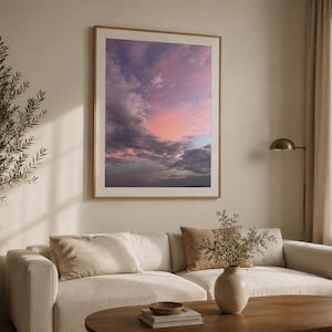 Peut inclure: Une impression d'art murale rectangulaire dans un cadre en bois clair représentant une photographie d'un ciel au coucher du soleil avec des nuages roses, violets et gris. L'œuvre est exposée sur un mur uni au-dessus d'un canapé beige.
