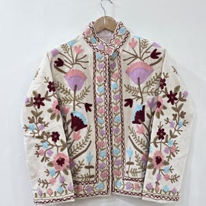 Pode incluir: Casaco bordado creme com gola alta. O casaco apresenta bordados florais coloridos em tons de rosa, roxo, azul e castanho. O bordado é detalhado e cobre a frente, a gola e as mangas do casaco.