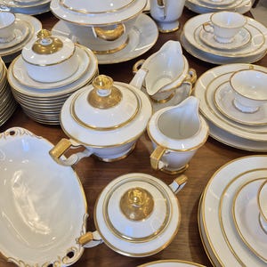 Op de afbeelding: Een grote witte porseleinen serviesset met gouden randen en accenten. De collectie bevat soepterrines met deksels, een koffiepot, een sauskom, een melkkan, diverse borden en theekopjes met schoteltjes, allemaal gerangschikt op een oppervlak.