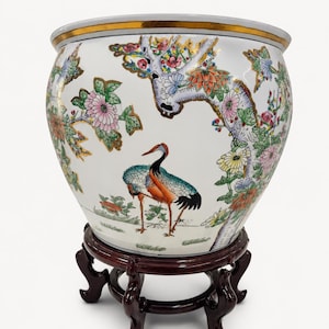 Peut inclure: Un cache-pot en céramique blanche orné d'un motif peint à la main représentant une grue parmi des fleurs colorées et des branches d'arbre avec des détails dorés. Le bol a un bord doré et repose sur un support en bois sculpté brun foncé.
