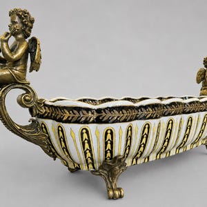 May include: An elongated ceramic centerpiece bowl featuring a white body with vertical black and yellow stripes. The bowl is supported by ornate bronze-toned metal feet and features two bronze-toned cherub figures at each end, one playing a flute.