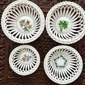 May include: A set of four white ceramic baskets with woven-style lattice sides and smooth circular bases. Each base features a unique floral or botanical illustration in green, pink, blue, and orange tones centered within the white ceramic.