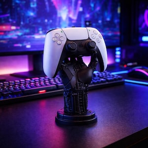 May include: A white and black gaming controller rests on a black controller stand. The stand is shaped like a hand. The background includes a keyboard, computer monitor, and mouse, all on a dark surface.