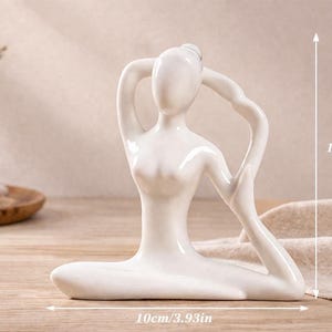 May include: A white ceramic yoga pose figurine, 4.33 inches tall and 3.93 inches wide, with arms raised overhead. The figurine is on a wooden surface, with a vase of greenery, a candle, and decorative stones in the background.