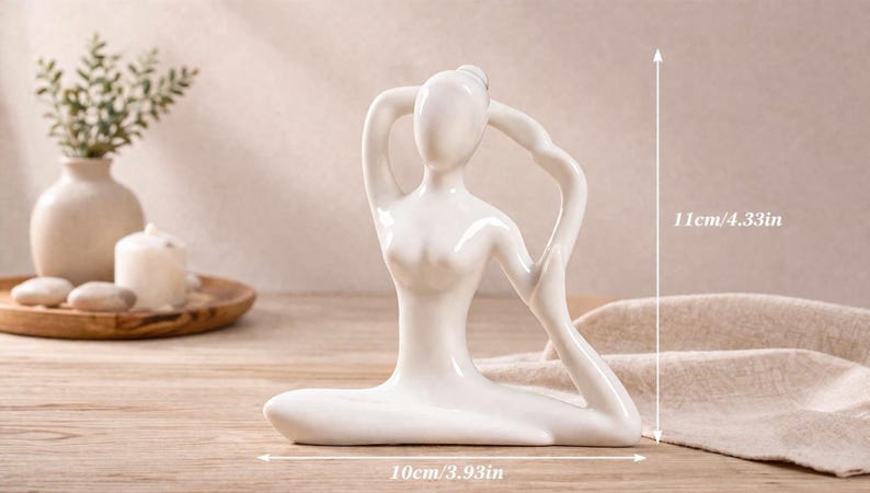 May include: A white ceramic yoga pose figurine, 4.33 inches tall and 3.93 inches wide, with arms raised overhead. The figurine is on a wooden surface, with a vase of greenery, a candle, and decorative stones in the background.