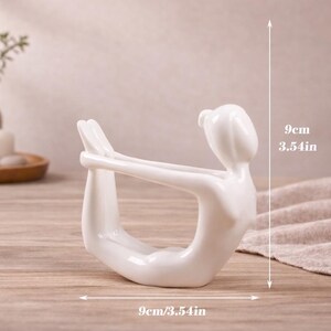 May include: A white ceramic yoga pose figurine, 3.54 inches tall and 3.54 inches wide, is displayed on a wooden surface. The figurine depicts a person in a yoga pose. The background includes a vase, candle, and decorative items.