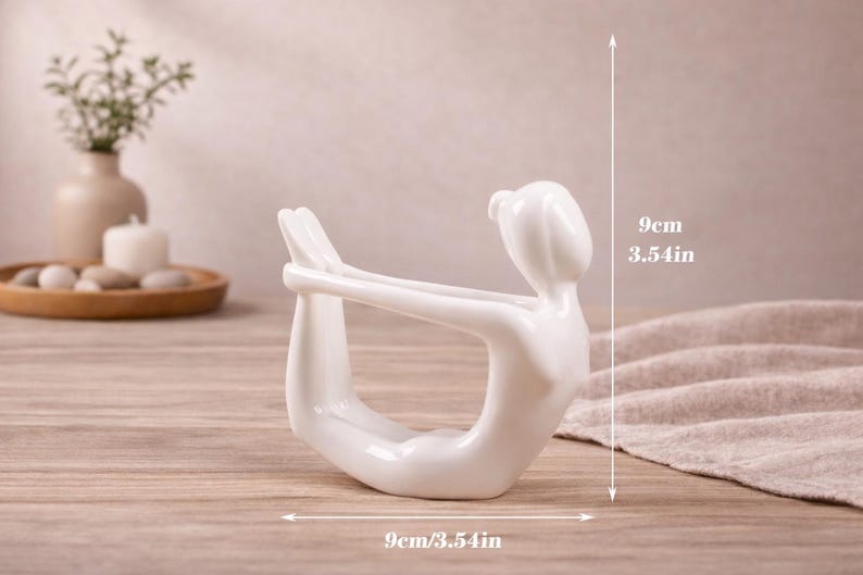 May include: A white ceramic yoga pose figurine, 3.54 inches tall and 3.54 inches wide, is displayed on a wooden surface. The figurine depicts a person in a yoga pose. The background includes a vase, candle, and decorative items.
