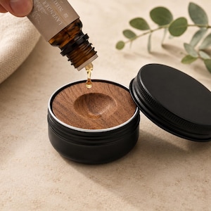 May include: A small black metal tin with a circular wooden insert featuring a central indentation. A single drop of amber liquid falls from a brown glass dropper bottle labeled Lavender into the wooden center of the open metal container.