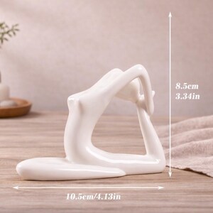 May include: A white ceramic yoga pose figurine. The sculpture is 4.13 inches long and 3.34 inches tall. The figurine is on a wooden surface. The background includes a vase with greenery, a candle, and decorative stones.