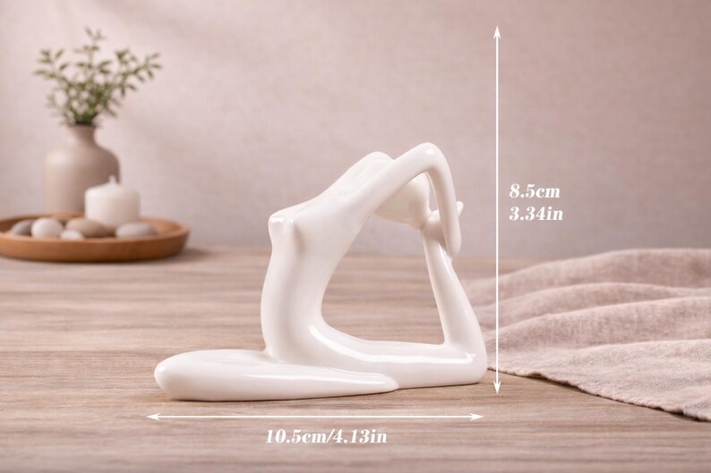 May include: A white ceramic yoga pose figurine. The sculpture is 4.13 inches long and 3.34 inches tall. The figurine is on a wooden surface. The background includes a vase with greenery, a candle, and decorative stones.