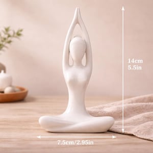 May include: A white ceramic yoga pose figurine. The figurine is 5.5 inches tall and 2.95 inches wide. The figure is in a seated lotus position with arms raised overhead. The background includes a wooden table and a decorative tray.
