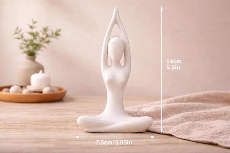 May include: A white ceramic yoga pose figurine. The figurine is 5.5 inches tall and 2.95 inches wide. The figure is in a seated lotus position with arms raised overhead. The background includes a wooden table and a decorative tray.