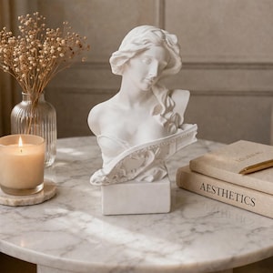 May include: A white marble bust of a woman with flowing hair sits on a marble table. A lit candle, a vase of dried flowers, and two books with the word "Aesthetics" are also on the table. A gold pen rests on the books.