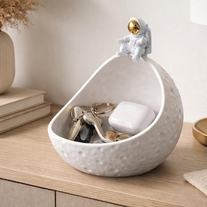 May include: A white, textured, moon-shaped decorative bowl with an astronaut figurine on top. The bowl contains keys and a white charging case. The astronaut has a gold-colored helmet. The bowl sits on a wooden surface.