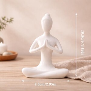 May include: A white ceramic yoga figurine in a seated pose with hands together in prayer. The figurine is 4.13 inches tall and 2.95 inches wide. The figurine is on a wooden surface.