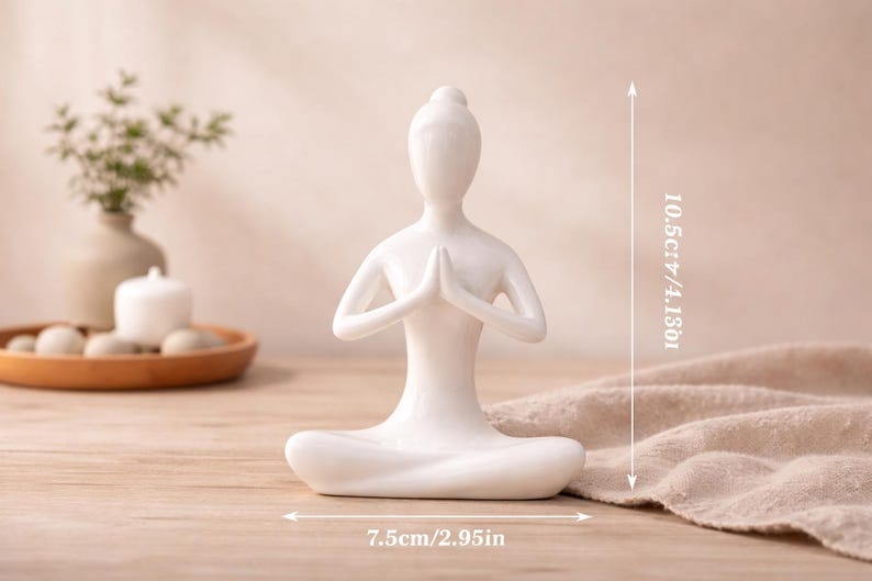 May include: A white ceramic yoga figurine in a seated pose with hands together in prayer. The figurine is 4.13 inches tall and 2.95 inches wide. The figurine is on a wooden surface.